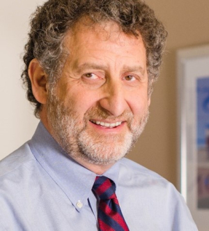 A person with short, curly brown and grey hair and a white beard smiles at the camera. He is wearing a dress shirt and tie.