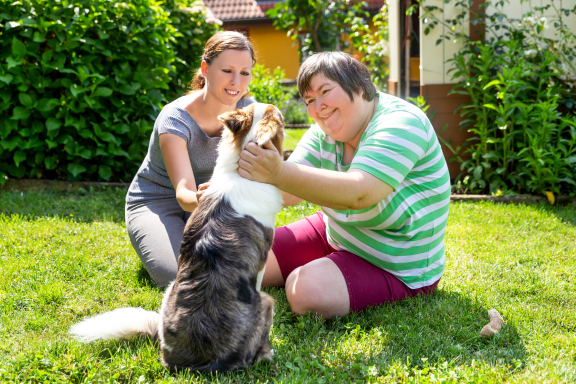 An adult with an adult caregiver pets a dog outside
