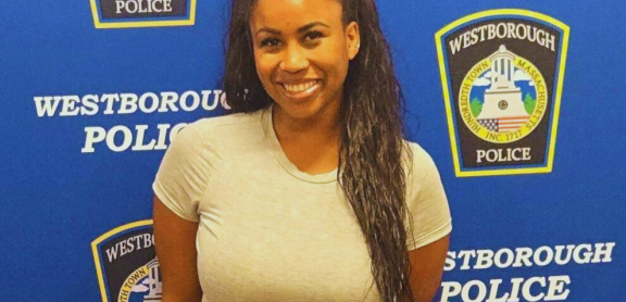 A smiling brown-skinned adult with long dark hair stands in front of a Westborough Police Department step-and-repeat background