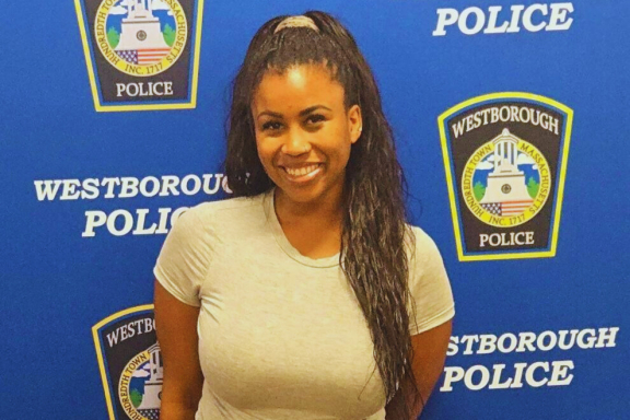 A smiling brown-skinned adult with long dark hair stands in front of a Westborough Police Department step-and-repeat background