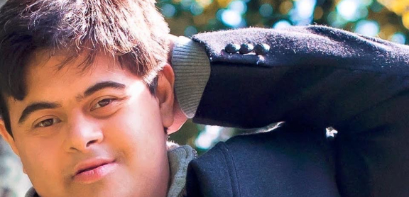 A person with short dark hair poses with his hand behind his head. He is outside on a sunny day.