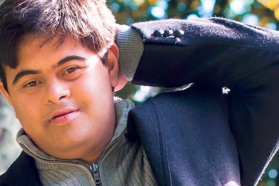 A person with short dark hair poses with his hand behind his head. He is outside on a sunny day.