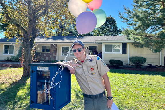 Eagle Scout Jim Kemp posing with one of the Little Free Libraries he and his troop installed at Advocates residential programs. There are colorful balloons tied to the library.