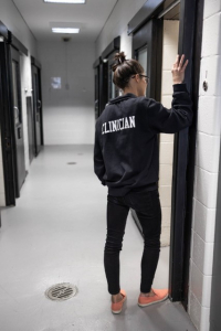 A clinician leans against a door frame outside a jail cell.