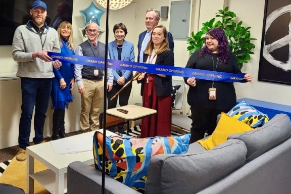 Seven adults in the VAULT space, preparing to cut a blue ceremonial ribbon